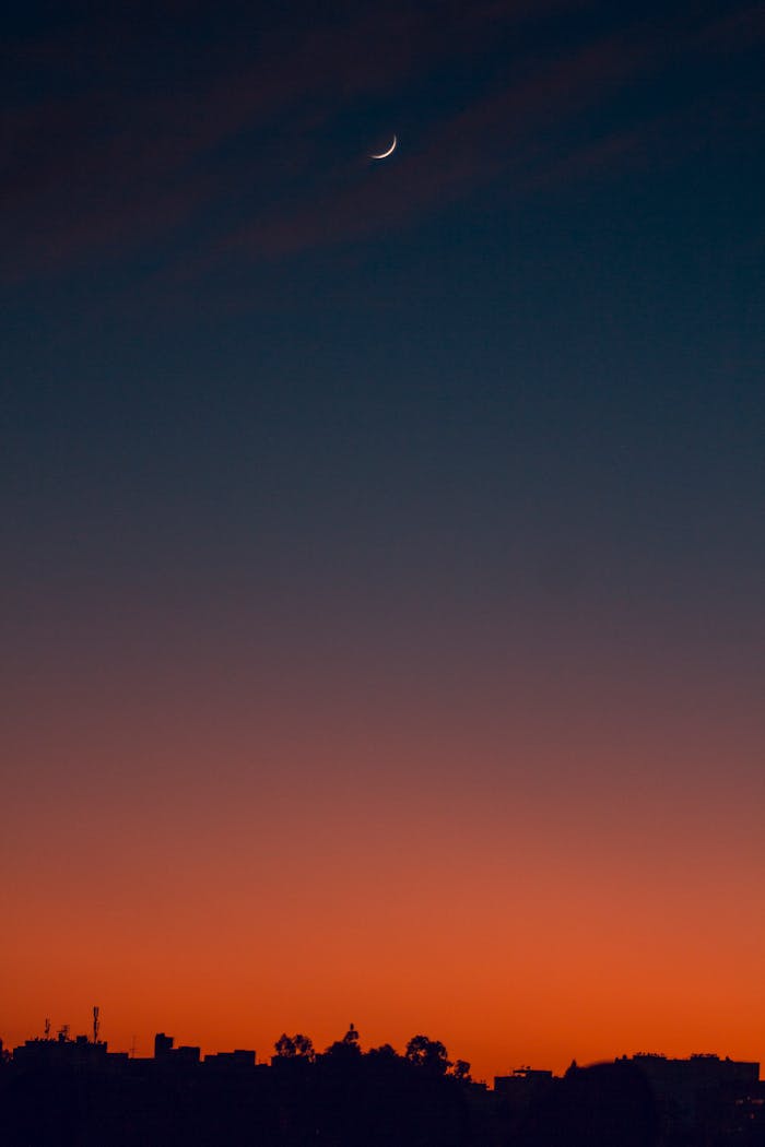 A serene crescent moon in a colorful dusk sky above a city silhouette, capturing twilight's beauty.